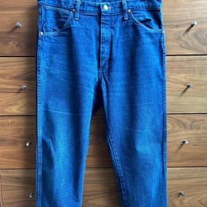 May include: A pair of classic blue denim jeans, hanging from a metal hanger. The jeans have a button and zipper closure, front and back pockets, and belt loops. The denim is a medium wash with subtle fading.