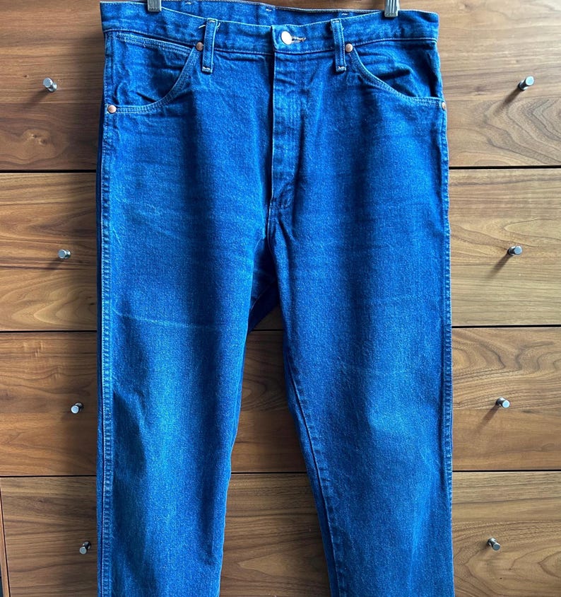 May include: A pair of classic blue denim jeans, hanging from a metal hanger. The jeans have a button and zipper closure, front and back pockets, and belt loops. The denim is a medium wash with subtle fading.