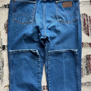 Pu&ograve; includere: Jeans blu in denim con un classico design a cinque tasche. Le tasche posteriori presentano un motivo di cucitura distintivo. Una toppa in pelle marrone con il marchio "Wrangler" &egrave; visibile sul retro. I jeans sono piegati e presentati.