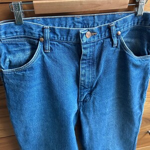 Pu&ograve; includere: Primo piano di jeans di denim blu con classico stile a cinque tasche. I jeans sono appesi a una gruccia di metallo. I jeans hanno una chiusura con bottoni e rivetti in rame. Il denim ha un lavaggio medio.