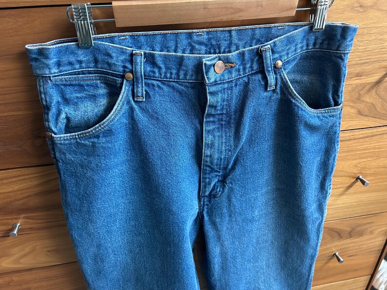 Pu&ograve; includere: Primo piano di jeans di denim blu con classico stile a cinque tasche. I jeans sono appesi a una gruccia di metallo. I jeans hanno una chiusura con bottoni e rivetti in rame. Il denim ha un lavaggio medio.