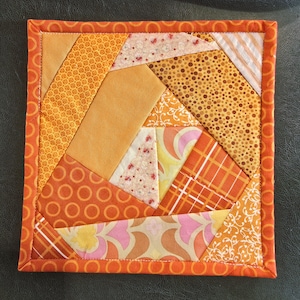 Puede incluir: Colcha de patchwork cuadrada en tonos naranja, amarillo y blanco. El diseño presenta formas geométricas y varios patrones, incluyendo lunares, estampados florales y rayas. Los bordes están acabados con un borde naranja liso.