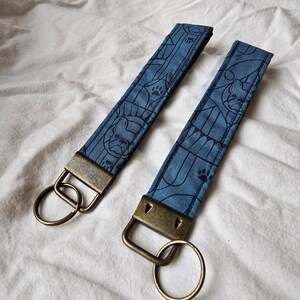 May include: Two blue fabric keychains with a pattern of French Bulldogs and paw prints. Each keychain has a brass key ring.