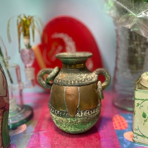 以下が含まれることがあります： 2つの取っ手のある、緑と銅色の小さなセラミックの花瓶。花瓶は表面がざらざらしていて、上部に銅の帯があります。