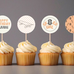 May include: Four cupcakes with white frosting topped with round paper cupcake toppers. The toppers feature a skateboard, the words "Happy Birthday Olivia!" and the words "Skater Girl" with the number 8. The fourth topper has a peach background with a pattern of lightning bolts and the number 8.