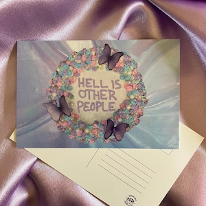 May include: A postcard featuring a cake design with the text "HELL IS OTHER PEOPLE." The cake is decorated with pastel frosting and butterfly accents. The postcard is on a lavender satin surface.