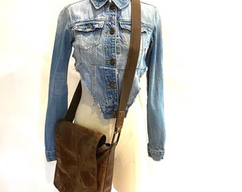 May include: A light blue denim jacket with a distressed hem and button closure. A brown leather messenger bag with a long strap hangs over the jacket. The bag has a flap closure and a worn appearance.