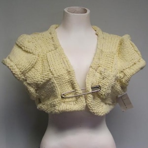May include: A pale yellow knitted shrug with a safety pin closure. The shrug has a V-neck and short sleeves.