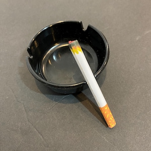May include: A black ceramic ashtray with a white cigarette resting inside. The cigarette is lit and has a red and orange ember.