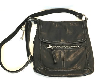 May include: Black leather crossbody bag with a flap closure and a front zippered pocket. The bag features white stitching and a long, adjustable strap. A silver-colored key charm hangs from the strap.