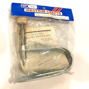 May include: A Westrim Crafts rhinestone setter tool, packaged in clear plastic. The tool includes a spring-loaded plunger, a metal base, and a wooden handle. The packaging features the text "LAURI SETTER" and "RHINESTONES SETTER".