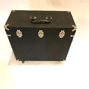 May include: A black trunk with silver metal corner protectors and clasps. The trunk has a black leather handle and sits on small wheels. The trunk is rectangular and appears to be made of wood or a similar material.