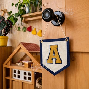 Puede incluir: Un banderín de fieltro crema con borde azul marino y la letra dorada "A" colgando de un pomo de puerta. El banderín tiene ojales dorados. Una casa de muñecas de madera y plantas en el fondo.