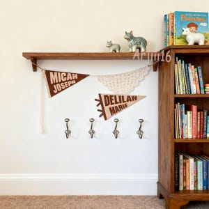 May include: A wooden shelf displays decorative items and pennant banners with names. Below, four metal hooks are mounted on the wall. A wooden bookcase filled with books is on the right.