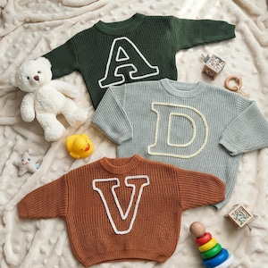 May include: Three knitted sweaters in green, gray, and brown, each featuring a large letter applique: A, D, and V. The sweaters are on a cream-colored blanket, with a teddy bear and other toys.