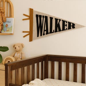 May include: A beige felt pennant with the name "WALKER" in black letters. The pennant has brown accents and is hanging on a white wall. A wooden crib is in the foreground, and a teddy bear is visible.