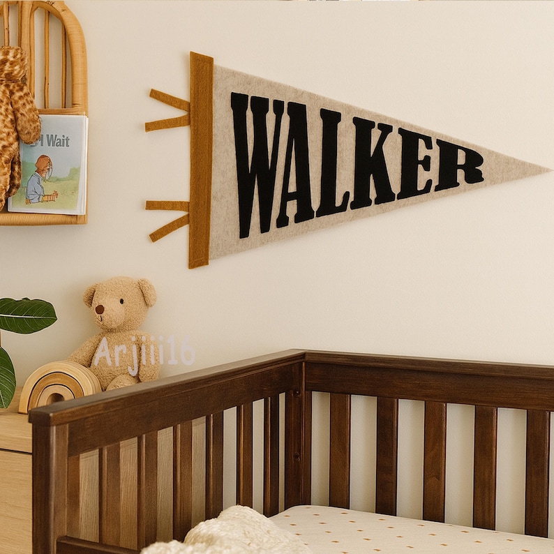 May include: A beige felt pennant with the name "WALKER" in black letters. The pennant has brown accents and is hanging on a white wall. A wooden crib is in the foreground, and a teddy bear is visible.