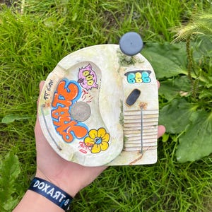 May include: A hand-painted, off-white ashtray with graffiti-style art. The design includes orange and blue lettering, a cartoon character, and a yellow flower. The ashtray has a small, gray knob and a vertical, textured section.