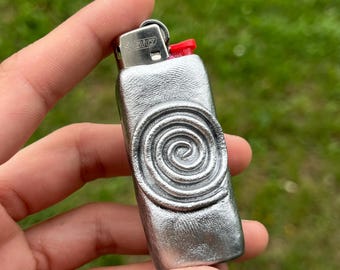 Puede incluir: Un encendedor de color plateado con un botón de encendido rojo. El encendedor tiene una superficie texturizada y un diseño en espiral en la parte delantera. El encendedor se sostiene en la mano de una persona contra un fondo verde borroso.