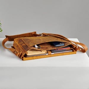 Könnte beinhalten: Eine braune Ledertasche mit einem Klappverschluss und einem langen verstellbaren Riemen. Die Tasche ist offen und mit verschiedenen Gegenständen gefüllt, darunter ein Telefon, Stifte und ein Notizbuch.