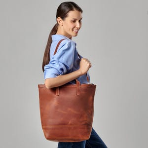 Könnte beinhalten: Eine braune Ledertasche mit Reißverschluss. Die Tasche hat einen oberen Griff und einen Schultergurt. Die Tasche wird von einer Person getragen, die ein hellblaues Hemd trägt.