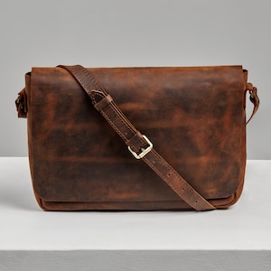 May include: A brown leather messenger bag with a long adjustable strap and a brass buckle. The bag has a distressed look and is made of high-quality leather.