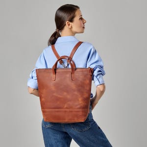 Puede incluir: Una mochila de cuero marrón con cierre de cremallera y correas ajustables. La mochila la lleva puesta una persona que lleva una camisa azul claro y unos vaqueros azules.