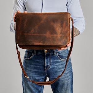 May include: A rectangular brown leather messenger bag with a large front flap, a long adjustable shoulder strap, and visible stitching along the edges. The leather has a distressed, matte finish with natural color variations throughout the surface.