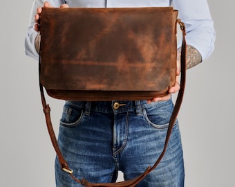 May include: A rectangular brown leather messenger bag with a large front flap, a long adjustable shoulder strap, and visible stitching along the edges. The leather has a distressed, matte finish with natural color variations throughout the surface.