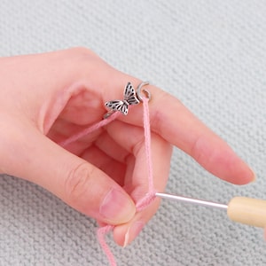 Puede incluir: Un anillo de mariposa plateado en un dedo, con hilo rosa que se está tejiendo a ganchillo. El anillo presenta un diseño detallado de mariposa. También se ve un ganchillo con mango de madera, junto con el hilo rosa.