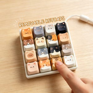 May include: A set of sixteen cat-themed keyboard keycaps in various colors, including orange, gray, and white. Each keycap features a different cat face design. The text "REMOVABLE KEYCAPS" is displayed above the keycaps.