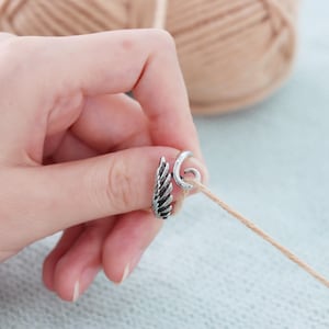 May include: A silver yarn guide ring featuring a feather design. The ring is wrapped around a strand of beige yarn, with a ball of yarn in the background. The ring is designed to help guide yarn while knitting or crocheting.