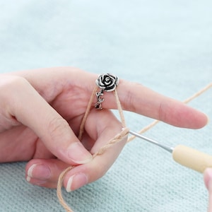 May include: A close-up of a hand using a crochet hook to work with tan yarn. A silver charm in the shape of a rose is on the index finger. The background is a light blue, textured surface.