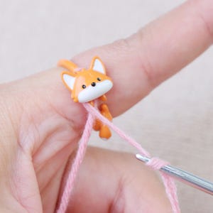 May include: An orange fox-shaped stitch marker with white accents is attached to pink yarn. The marker is on a finger, with a crochet hook in the yarn. The fox has black eyes and a white muzzle.