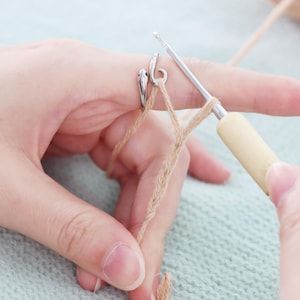 May include: Close-up of a crochet project in progress. A silver yarn guide ring is on a finger, with beige yarn being worked with a crochet hook. The hook has a light-colored wooden handle. The background is a light green knitted fabric.
