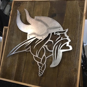 May include: A metal cutout of a Viking helmet on a wooden background. The helmet is silver and has a detailed design. The background is made of wood planks.