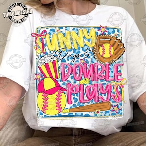 May include: White t-shirt featuring a colorful graphic design with the text "Sunny Days & Double Plays". The design includes a baseball glove, baseball, popcorn, a baseball cap, and a baseball bat. The design is in yellow, pink, and blue.
