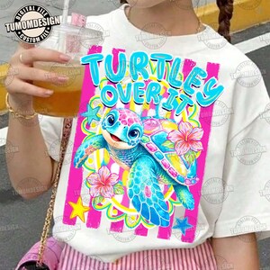May include: White t-shirt featuring a colorful graphic of a cartoon turtle with the text "TURTLES OVER IT". The turtle has a pink and blue shell, blue flippers, and is surrounded by flowers and stars. The design is set against a pink striped background.