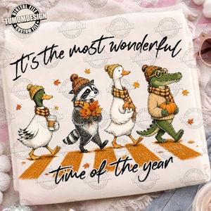 May include: A white t-shirt featuring a graphic of a duck, a raccoon, a goose, and an alligator wearing autumn hats and scarves. The text reads, It's the most wonderful time of the year. The animals are walking on a yellow crosswalk with falling leaves.