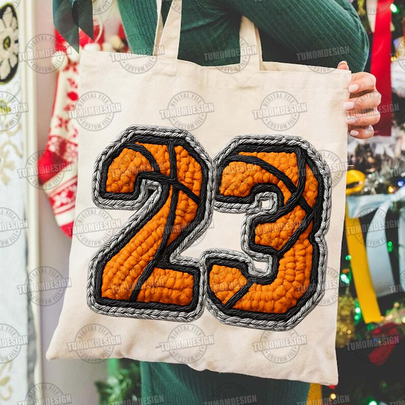 May include: A cream tote bag displaying the number "23" in an orange basketball design with black lines and a grey stitched border. The bag is held by a person. The design is reminiscent of a basketball.