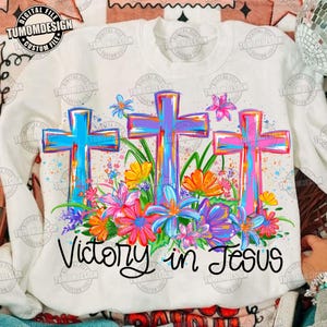 May include: White sweatshirt featuring three colorful crosses with a floral arrangement and the text "Victory in Jesus." The crosses are painted in shades of blue, purple, and pink, with a variety of flowers in vibrant colors.