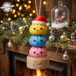 May include: A hanging Christmas ornament featuring a stack of three round, smiling characters in yellow, blue, and pink, topped with a red Santa hat. The stack rests inside a small, light-coloured wooden bamboo steamer basket with twine.