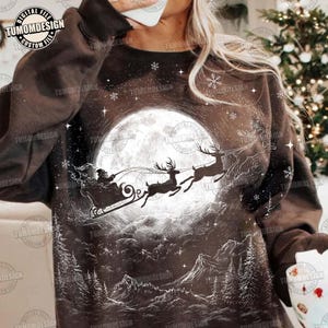 May include: A dark brown sweatshirt featuring a black and white graphic of Santa Claus in a sleigh pulled by reindeer against a full moon. The design includes a snowy landscape and the text "TUMOMDESIGN".