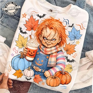 Puede incluir: Una sudadera blanca con el gráfico de una muñeca de pelo naranja, que viste un peto azul y una camiseta a rayas. La muñeca sostiene una taza con una cara de calabaza. El diseño incluye hojas de otoño de colores, calabazas y murciélagos.
