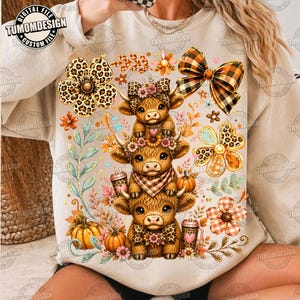 May include: A beige crewneck sweatshirt featuring a graphic print of three stacked Highland cows. The design includes leopard print flowers, a plaid bow, pumpkins, and autumn foliage. The text TUMOMDESIGN appears in the top left corner.