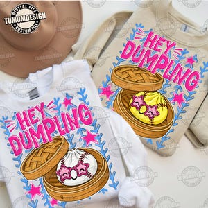May include: Two long-sleeved sweatshirts, one white and one beige, feature the text "HEY DUMPLING" in pink. Each shirt displays a cartoon dumpling with star-shaped sunglasses inside a bamboo steamer, surrounded by pink stars and blue accents.