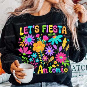 Op de afbeelding: Zwarte sweatshirt met een kleurrijk bloemmotief en de tekst "LET'S FIESTA" in heldere letters. Daaronder de naam "Antonio" in een levendig lettertype, omringd door bloemen in roze, gele en blauwe tinten.