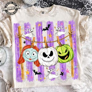 May include: A cream-colored t-shirt featuring a graphic of three pumpkins hanging from a clothesline against a purple striped background. The pumpkins resemble Sally, Jack Skellington, and Oogie Boogie, accompanied by ghost and bat illustrations.