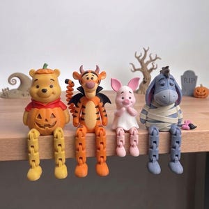 May include: Four articulated figurines of Winnie the Pooh characters sitting on a ledge. Pooh wears a pumpkin costume, Tigger is dressed as a devil with wings, Piglet wears a white ghost sheet, and Eeyore is wrapped in white bandages like a mummy.
