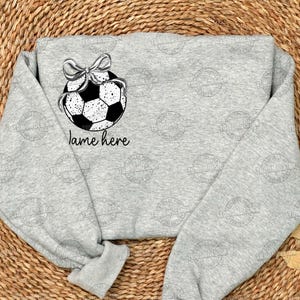 May include: A gray sweatshirt with a black and white soccer ball with a bow on top. The text "name here" is printed below the soccer ball.